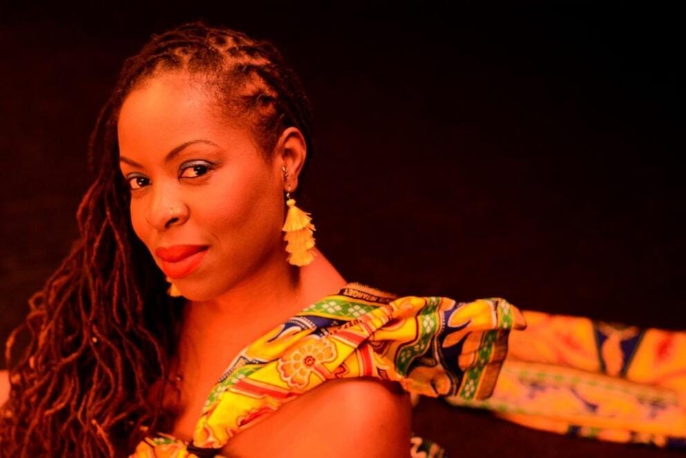 a Black woman with braids looks at the camera, a colorful scarf flies off the back of her shoulder