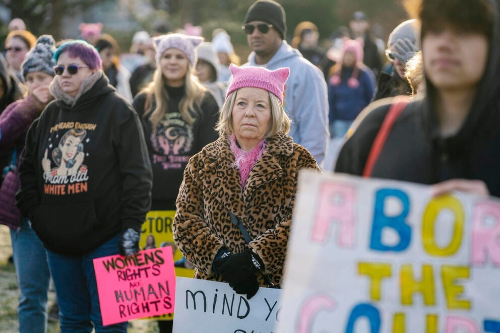 a woman in a pink hat stands in a crowd of protestors