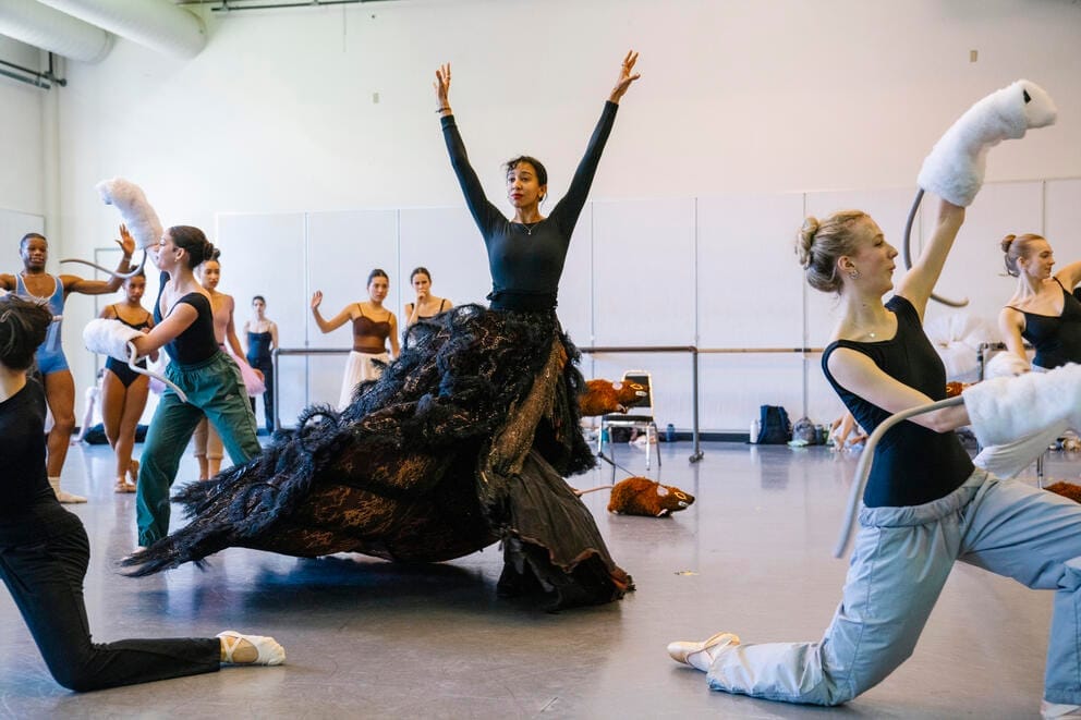 a ballet studio rehearsal scene with one woman in a black bodysuit and huge lacy skirt at center, arms upraised