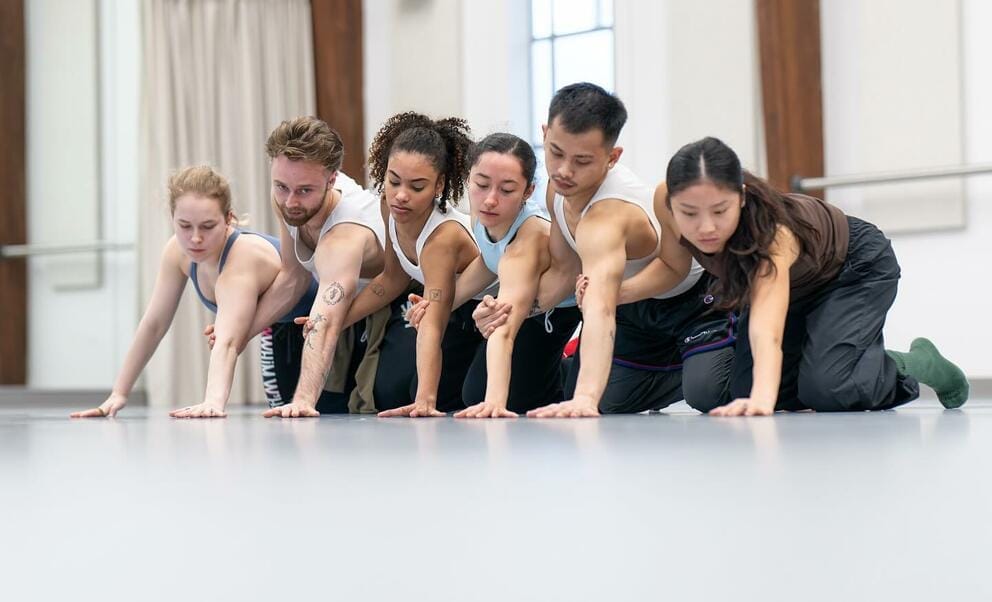 a row of dancers wearing white tanks and black pants kneels in a rehearsal studio