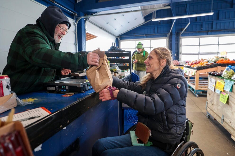 a woman in a wheelchair grabs a bag of groceries from a man working as a cashier