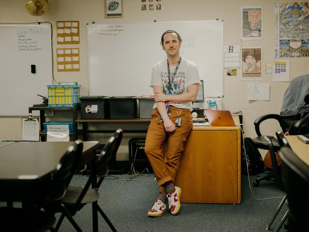 a teacher sits on a desk, looking at the camera