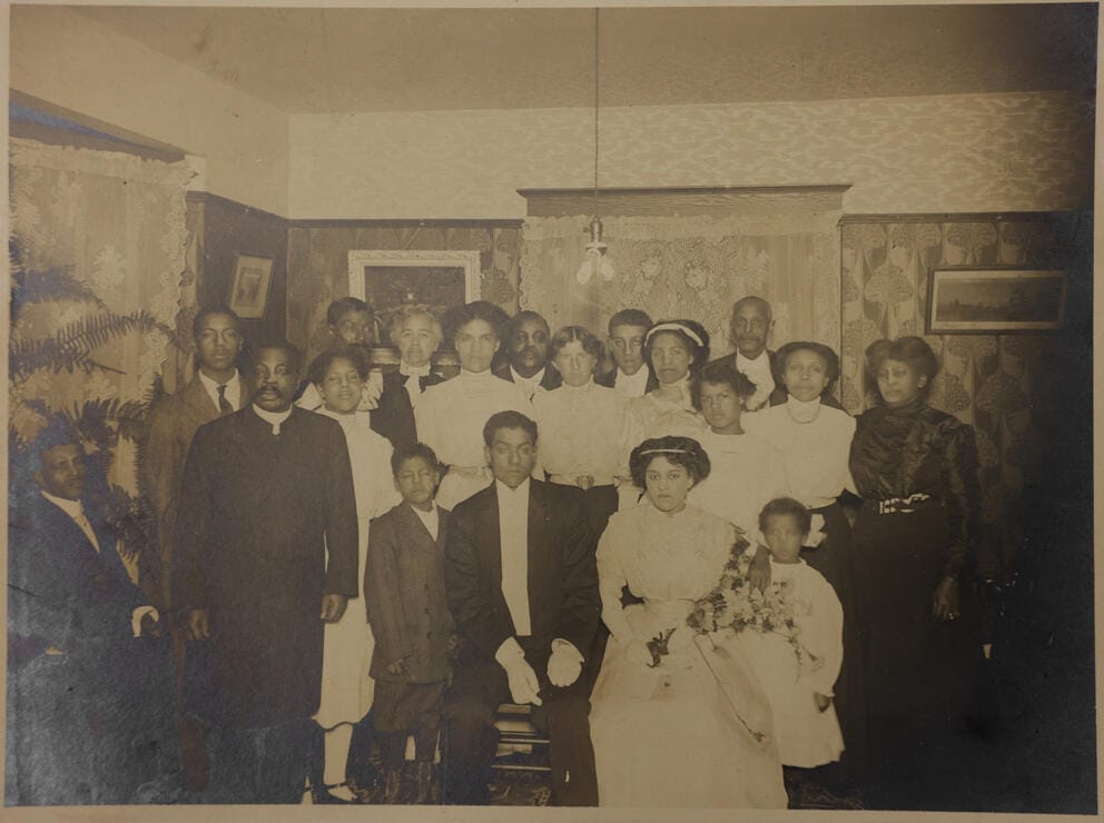 An old black and white photograph of a family during a wedding, the bride and groom are seated in front and their friends and family surround them