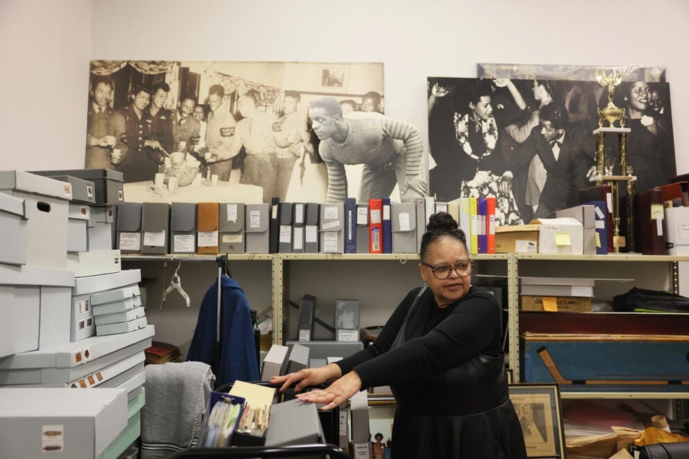A woman stands with her hands on collections boxes in a room filled with archival items, enlarged photographs line the wall behind her