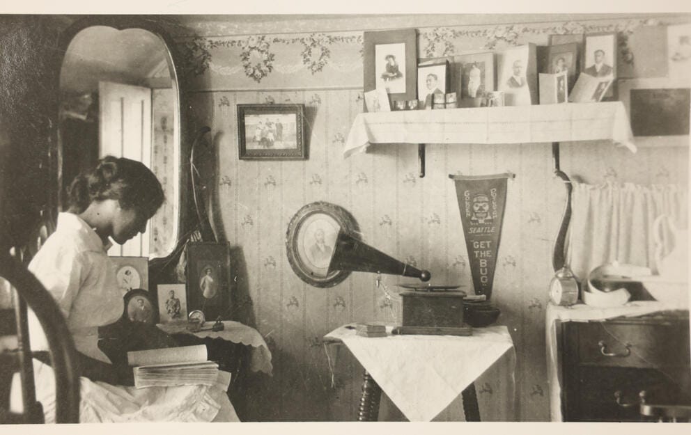 A black and white photograph of a woman sitting in a chair in a bedroom, photographs, a pennant and a phonograph line the walls