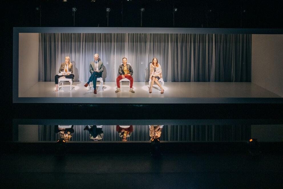 a horizontal-box stage set with four actors sitting on chairs inside it