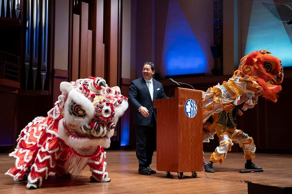 two dancers dressed in Chinese lion costumes perform on stage on either side of a man in a dark blue suit
