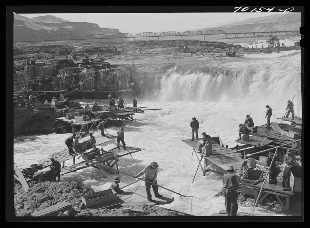 a black and white photograph showing people fishing in a river near a natural waterfall.
