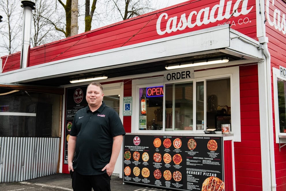 Thomas Reinhard stands outside a the storefront of Cascadia Pizza. A menu with pizzas is visible in the background.