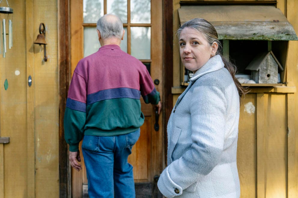 A person faces the camera, while an older person's back is to the camera.