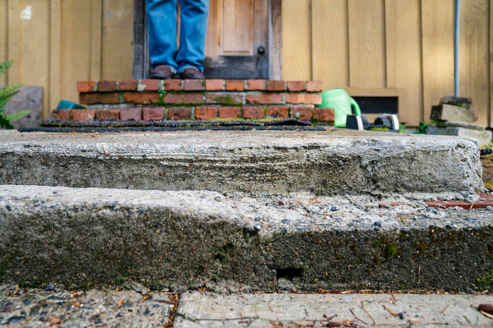 Dilapidated and uneven concrete steps in the foreground. Some distance back, someone is seen from the knees down to their feet.