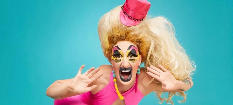 photo of a person in exaggerated female drag, with wildly piled blond hair and insanely garish eye makeup