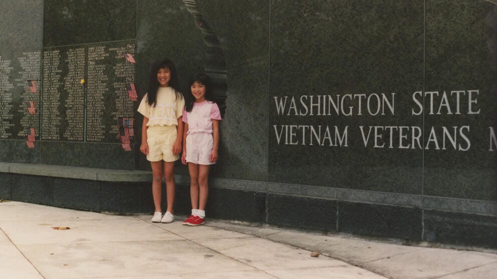 Two young girls stand in front of a large black marble wall with words inscribed on it.
