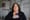 A tight portrait of Renee Raketty. She is wearing black and holding a U.S. passport over heart. There is an LGBT pride flag hanging in the background.