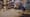 An elder man with long white hair and a beard works at a table in a room crowded with art supplies.