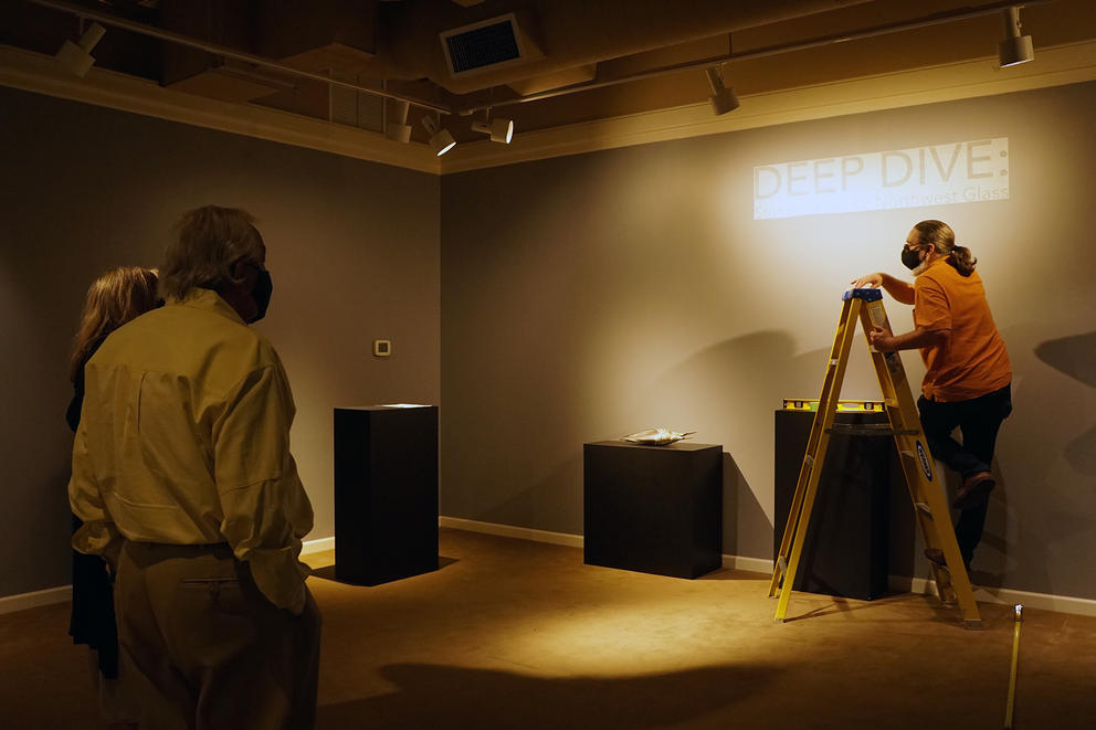 Person on ladder hangs up art while two onlookers stand by
