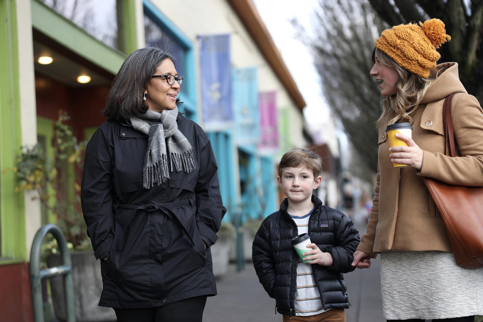 Marilyn Strickland talks to a woman and a child on a sidewalk, with colorful storefronts in background