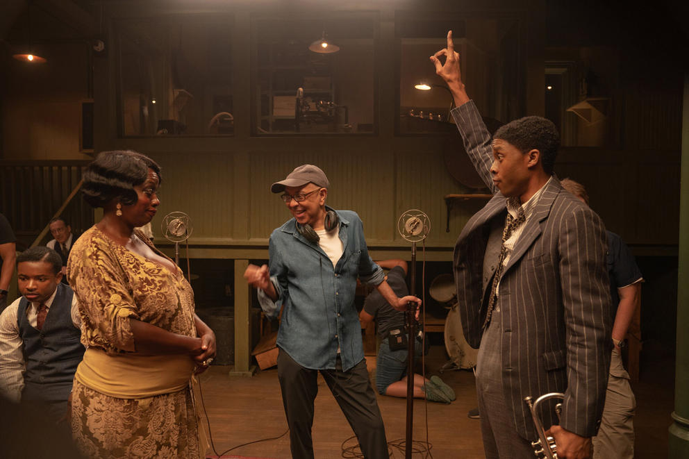 two actors in jazz era clothes, one with arm raised, the other in yellow dress, looking at film director in the middle