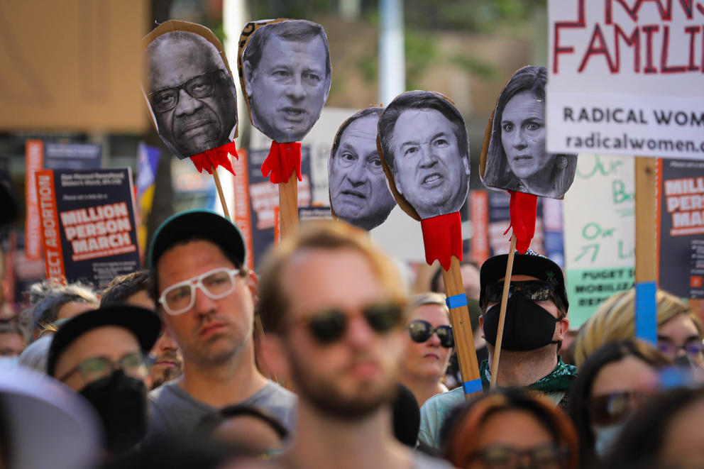 cutouts of five faces are held above a crowd of people