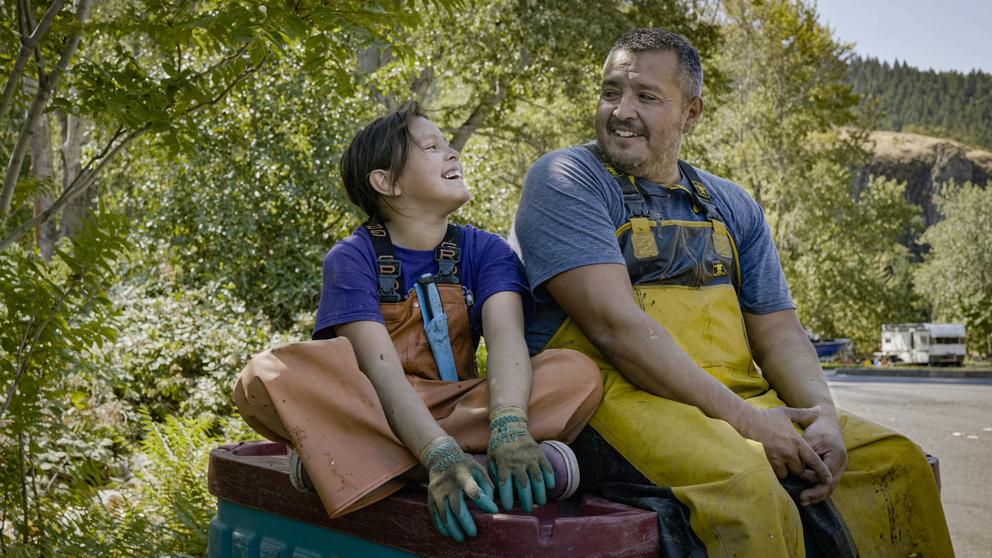 A young girl sits cross-legged while smiling up at her father sitting next to her. Both wear fishing waders.