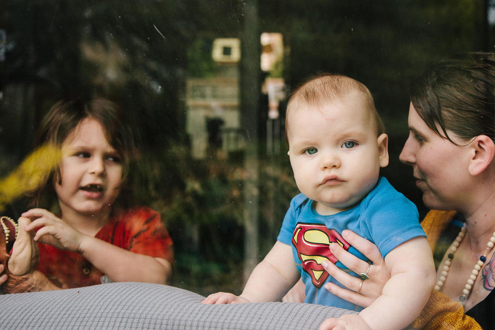 A baby looks at the camera while held up to the window by their mother