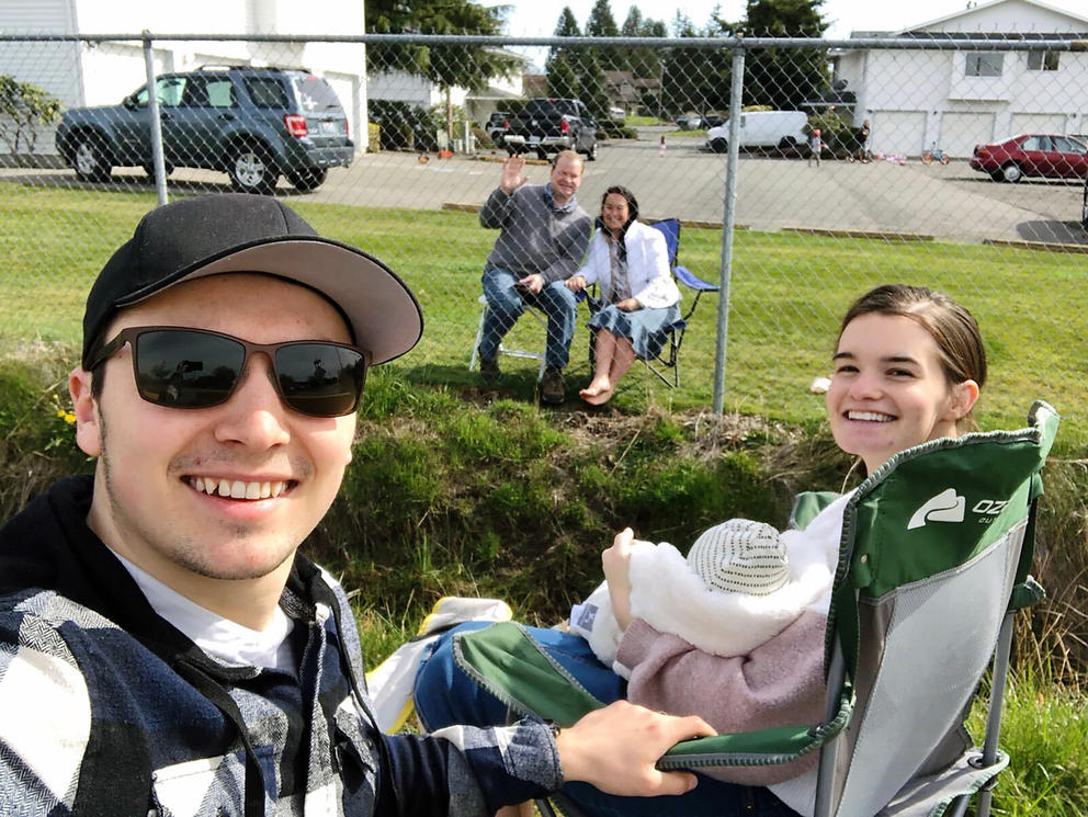 A young couple with a baby sit in camp chairs while grandparents sit on the other side of a chainlink fence