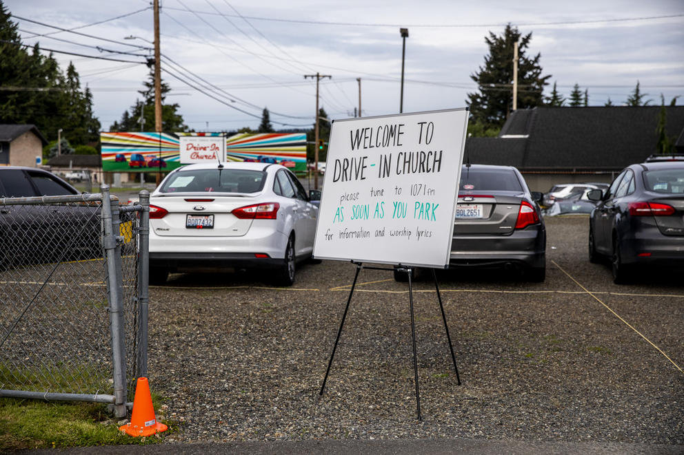 A whiteboard outside a parking lot reading "Welcome to drive-in church. Please tune to 107.1 FM for information and worship lyrics"