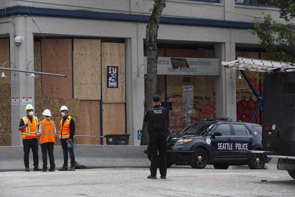 a police officer stands in middle of street, looks at a boarded up precinct with a Black Lives Matter poster on it
