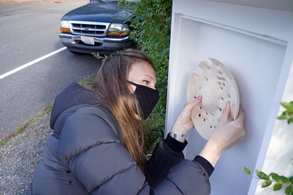 Woman in black coat attaches a wooden, carved disk to a white box