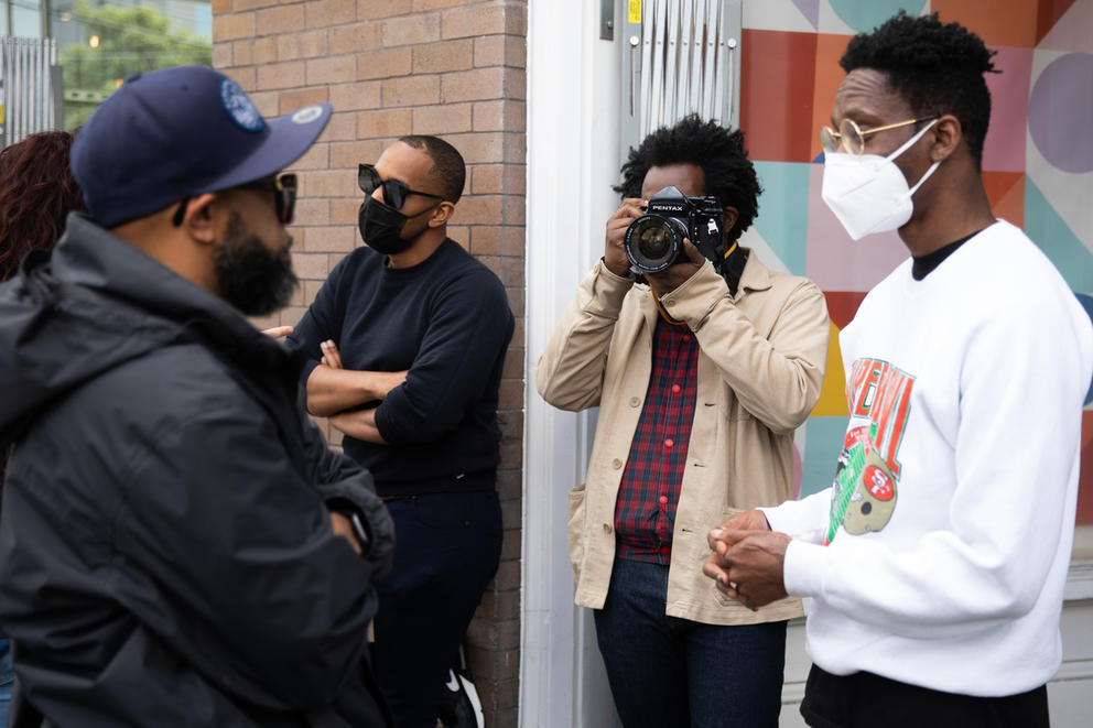 A man standing near a brick storefront takes a photo while two men talk in front of him