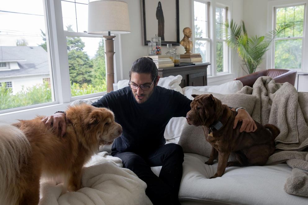 Abraham Dairi sits on a couch with his two dogs, windows behind him