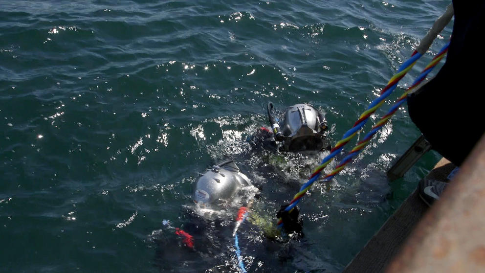 Two divers wearing metal helmets descent into the ocean