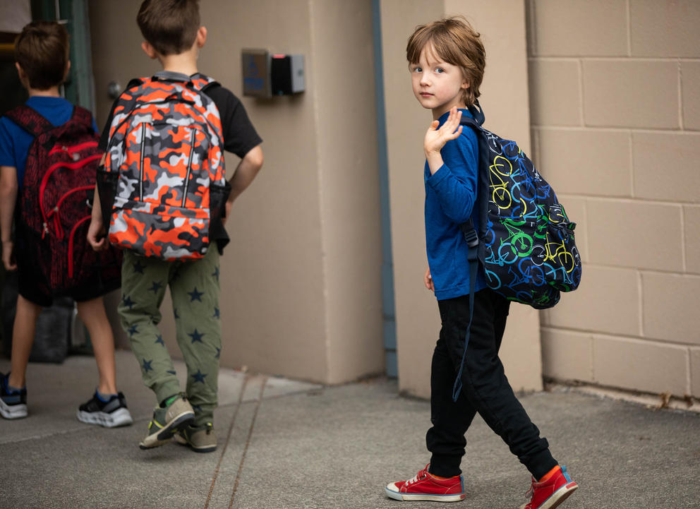 A young student wearing a backpack turns to wave while walking through a school door.