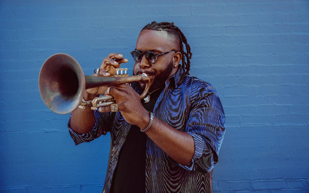 Person in blue shirt plays the trumpet in front of a blue wall 