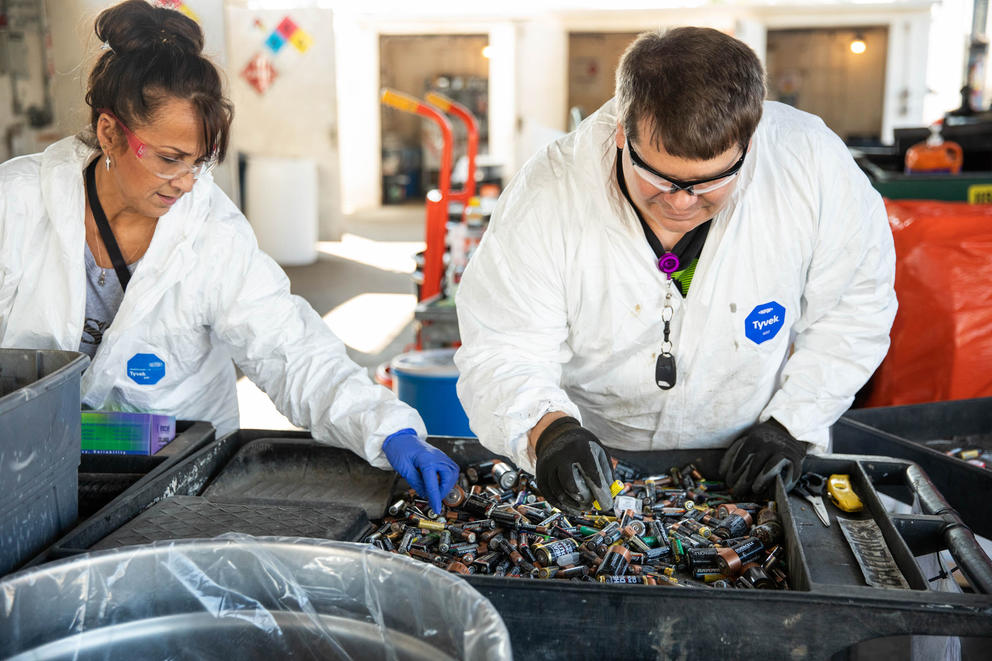 two people in white coats and protective glasses sort batteries 