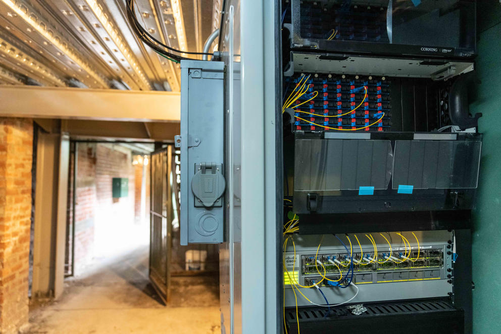 A gray panel box of fiber inputs and connections hangs on a wall in a tunnel.