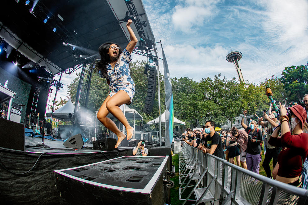 Person in blue and white clothing jumps on a stage, crowd is to the right, blue sky and space needle in the upper right corner