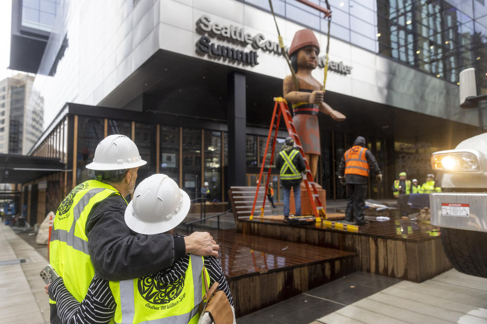 Artist Andrea M. Wilbur-Sigo and husband Steve Sigo wear protective equipment and embrace as workers install “Mowitch Man,” one of the two Salish welcome figures she carved. (Jason Redmond for Crosscut)