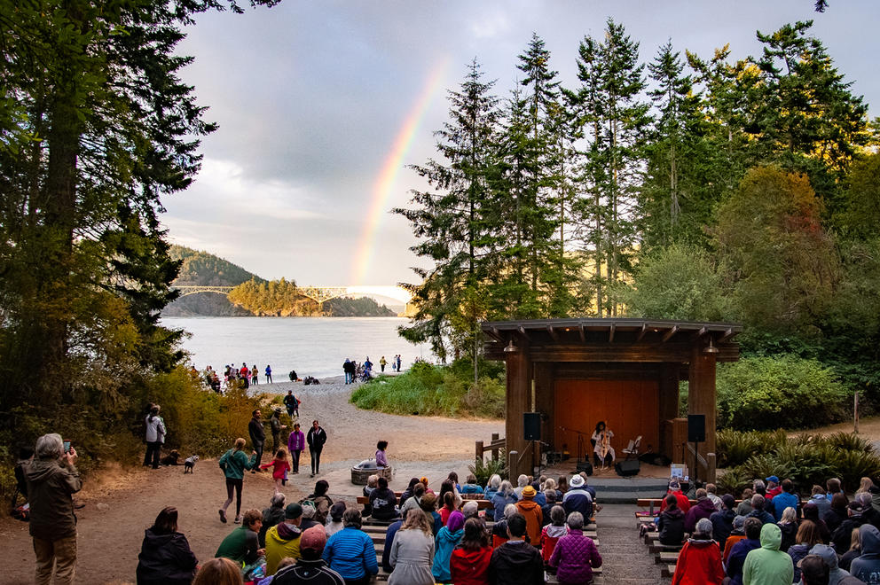A covered stage features musicians, a crowd sits in front. In the back: trees, water, and a rainbow.