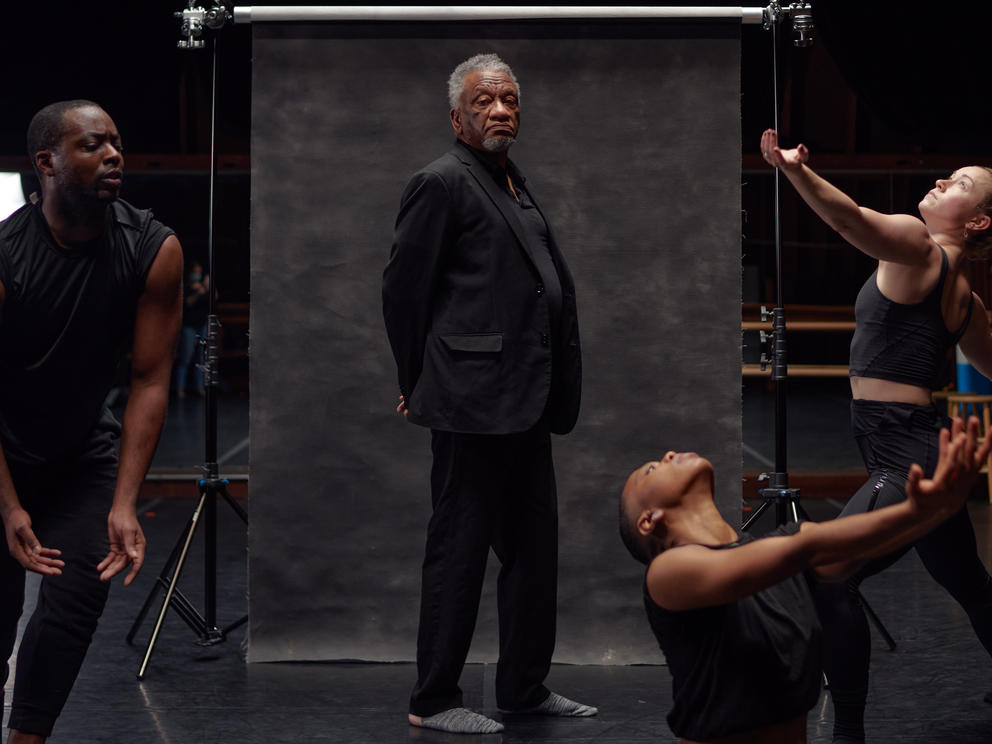 A person in a black suit with hands folded behind his back stands in front of a dark screen, as dancers move around him