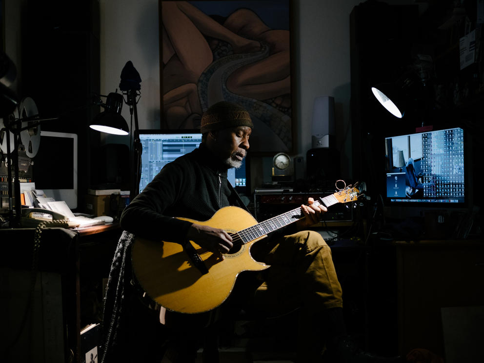 Person in studio with guitar lit by a light and blue screens behind 