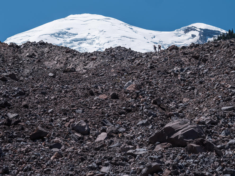 Two people walking on a rocky slope with a snowy peak behind them