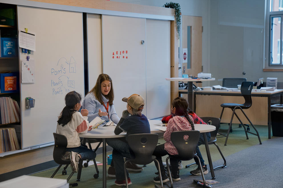 An education professional talks while sitting at a small table with four young students who are reading books.