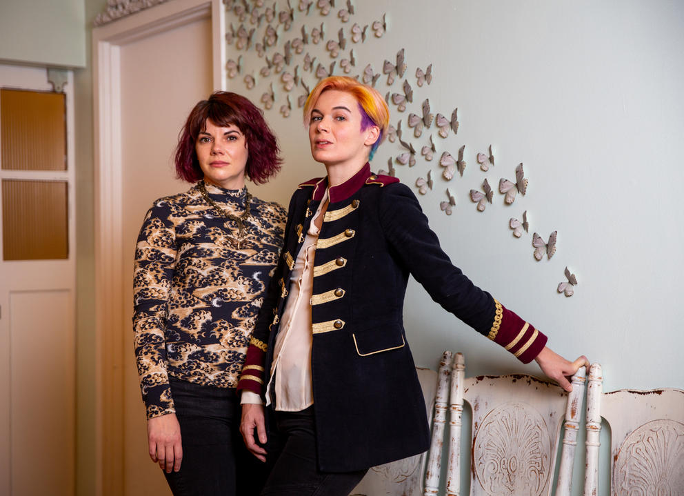 Two people standing in front of a mint-tinted wall featuring a swarm of white butterflies. Both people are dressed in shades of beige, black and white and have short hair.