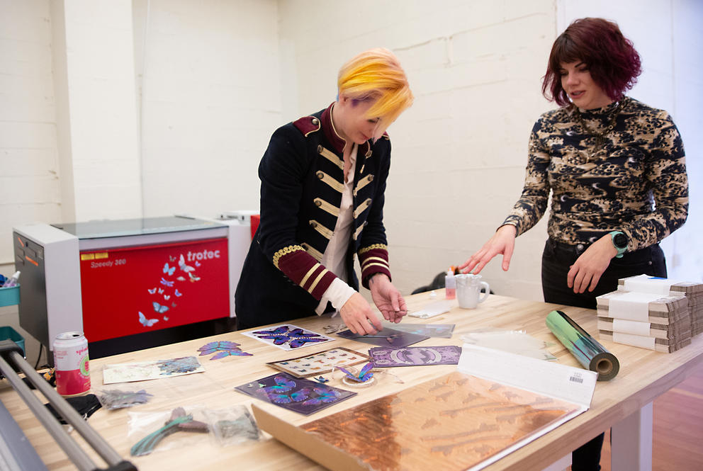Two people standing at a large table, with a printer in the background. They are holding paper butterflies.