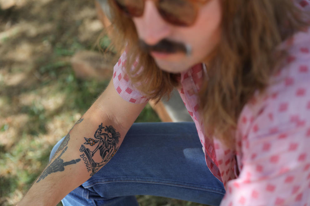 Person in pink shirt with mustache and sunglasses shows their arm with tattoos