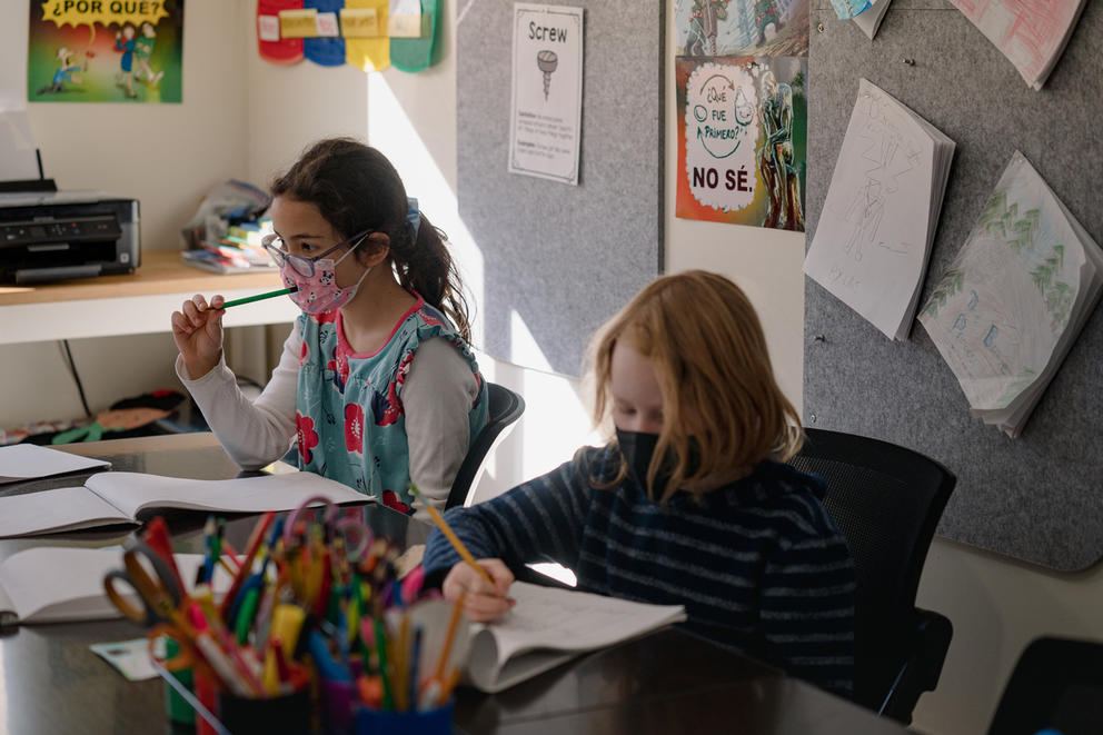 Two young students sit at a desk writing and drawing for an assignment.