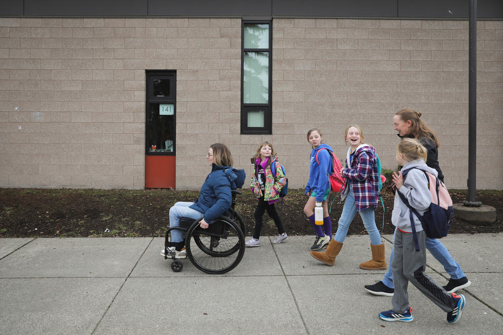 Four kids and two women walk on a sidewalk near a school.