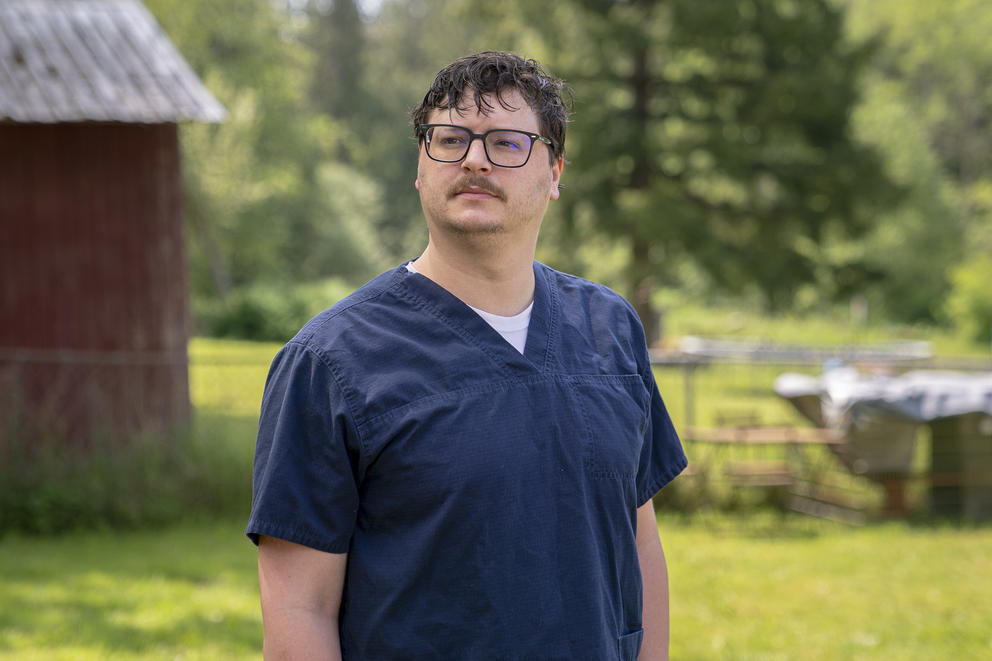 Trevor Gjendem poses in dark blue scrubs in front of a wooded area. 