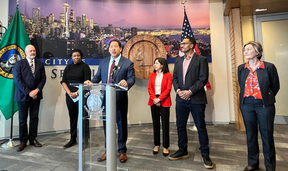 the seattle mayor and five councilmembers stand at a lecturn in city hall